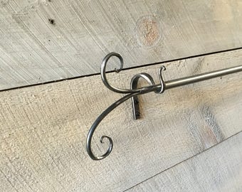 May include: A handcrafted metal curtain rod with decorative scrollwork on the ends. The rod is mounted on a light-colored, rustic wooden wall. The metal has a brushed, matte finish, adding a touch of elegance to the design.