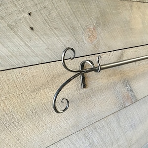 May include: A handcrafted metal curtain rod with decorative scrollwork on the ends. The rod is mounted on a light-colored, rustic wooden wall. The metal has a brushed, matte finish, adding a touch of elegance to the design.