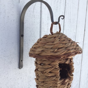 May include: A woven birdhouse with a natural brown color hangs from a metal hook. The birdhouse has a round roof and a square opening for birds to enter.