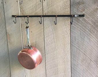 May include: A black metal pot rack with five hooks, mounted on a wood wall. A copper pot hangs from one of the hooks.