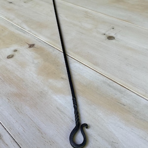 May include: A black, hand-forged fireplace poker with a U-shaped end and a twisted handle. The poker has a curved hook at the bottom. The tool is designed for tending to a fire.