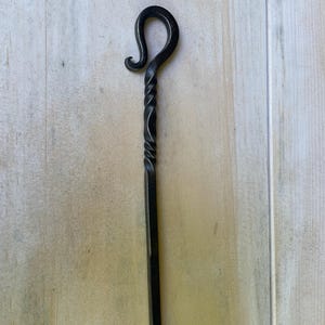 May include: A hand-forged, black metal fire poker with a twisted shaft and a curled hook at the top. The poker is designed for use in a fireplace or fire pit. The metal has a slightly textured surface.