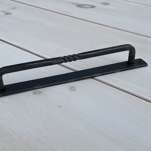 May include: Black metal drawer pull with a twisted design. The pull is mounted on a rectangular base.