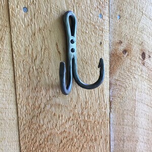 May include: A black metal hook with a double hook design. The hook is mounted on a wooden wall.