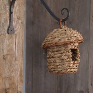 May include: A woven birdhouse with a thatched roof hangs from a black metal hook attached to a wooden wall.