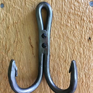 May include: A double hook made of metal with a hammered finish. The hook has a loop at the top and two curved hooks at the bottom. The hook is mounted on a wooden surface.