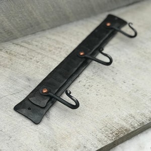 以下が含まれることがあります： 3つのフックと銅のアクセントが付いた黒い金属製の壁掛けフックラック。ラックは薄い灰色の木製の表面に取り付けられています。
