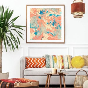 May include: A white couch with orange, blue, and white patterned throw pillows. A yellow throw pillow is on the right side of the couch. A wooden coffee table with a candle and a vase is in front of the couch. A framed print of a marbled design in shades of blue, pink, and yellow hangs on the wall behind the couch.