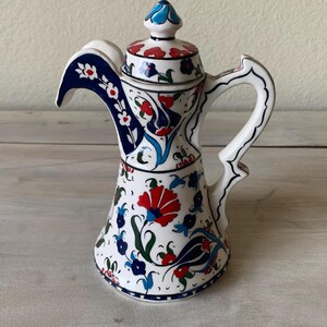 May include: White ceramic pitcher with a lid, adorned with a vibrant floral pattern in red, blue, and green. The pitcher features a curved spout and handle, showcasing a traditional design. The lid is topped with a blue finial.