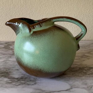 May include: A round ceramic pitcher featuring a two-tone glaze with a light mint green center that transitions into a dark brown finish at the base and rim. The pitcher includes a curved handle and a molded pouring spout on the upper edge.