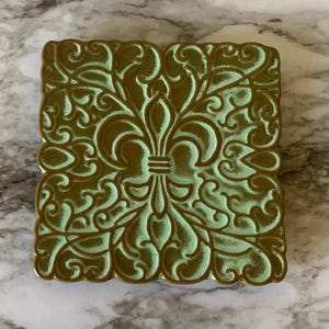 May include: A square decorative tile featuring a central fleur-de-lis emblem surrounded by intricate scrolling filigree patterns. The design displays a two-tone color scheme with a light mint green background and raised bronze-colored detailing.