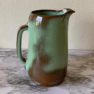 May include: A ceramic pitcher featuring a two-tone glaze with a glossy brown base that transitions into a speckled mint green upper section. The vessel includes a curved handle on the side and a prominent pouring spout at the top rim.