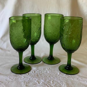 May include: A set of four green glass goblets featuring a distinct crackle texture on the bowls. Each goblet has a smooth stem and a circular base, standing upright. The glass is a consistent deep green color throughout the entire structure.