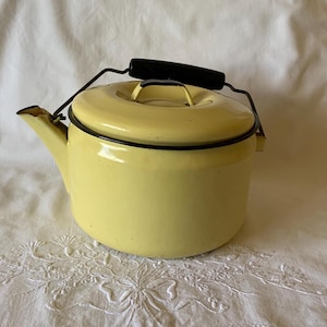 May include: A pale yellow enamel tea kettle with a black handle and a matching lid. The kettle features a dark rim around the top edge and a spout on the left side. The metal handle is hinged and includes a black grip for handling the vessel.