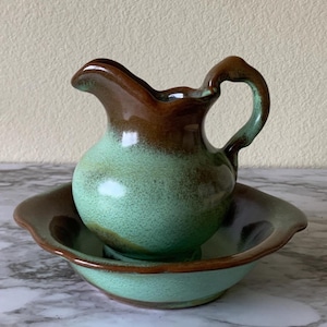 May include: A ceramic pitcher and basin set featuring a two-tone glaze with a speckled mint green body and a dark brown rim. The pitcher has a curved handle and a pouring spout, while the basin has a wide, scalloped edge and matching colors.