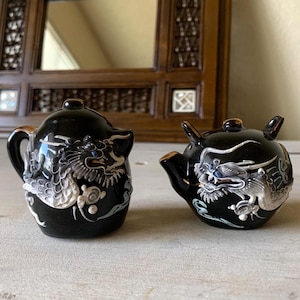 May include: Two black ceramic teapots with dragon designs. The dragons are detailed in silver and white, with blue accents. The teapots have gold trim and are set against a neutral background.