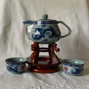 May include: A ceramic teapot with a lid and two matching cups, all featuring a blue and red dragon pattern on a light gray speckled background. The teapot rests on a dark brown wooden stand with a circular base and carved upright supports.