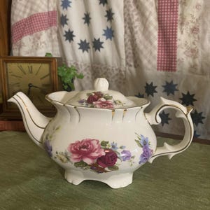 May include: A white porcelain teapot with a floral design featuring pink and purple roses and blue flowers. The teapot has a gold trim and a matching lid. The handle is curved and ornate. The teapot is on a green surface.