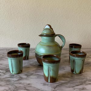 May include: A ceramic beverage set consisting of a lidded pitcher and four matching cups. Each piece features a speckled light green glaze with dark brown accents around the rims and bases. The pitcher has a rounded body and a curved handle.