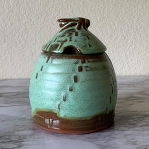 May include: A ceramic honey pot with a lid, featuring a light mint green glaze with speckled brown accents and a dark brown base. The lid has a small knob handle and a cutout for a dipper. The body has a rounded shape with horizontal ridges.