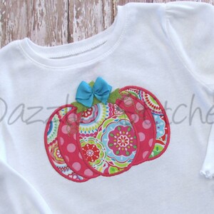May include: White long-sleeved shirt with a pink, green and blue patchwork pumpkin applique. The pumpkin has a blue bow on top.