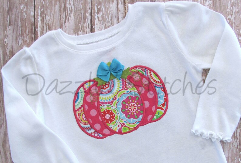 May include: White long-sleeved shirt with a pink, green and blue patchwork pumpkin applique. The pumpkin has a blue bow on top.
