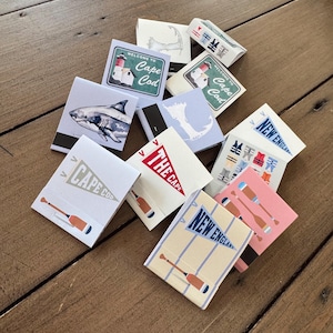 May include: A collection of rectangular matchboxes featuring various designs including a shark, lighthouses with the text Welcome to Cape Cod, pennants labeled Cape Cod and New England, oars, and nautical icons in blue, red, white, and pink color palettes.