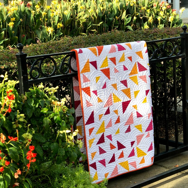 May include: A white quilt with a geometric pattern of red, orange, and yellow triangles. The quilt is hanging on a black metal fence in front of a bed of green plants.