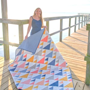 May include: A geometric quilt featuring a pattern of colorful triangles in shades of blue, coral, yellow, and gray. The quilt is displayed outdoors, showcasing its design and craftsmanship. The quilt's reverse side is a solid light blue.