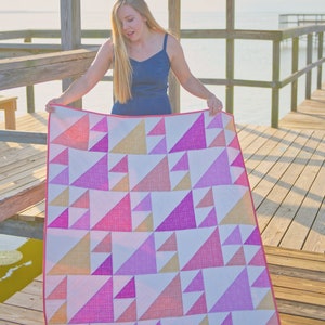 May include: A colorful quilt with a geometric pattern of triangles in shades of pink, orange, and white. The quilt has a pink border and is held up by a person on a wooden dock.