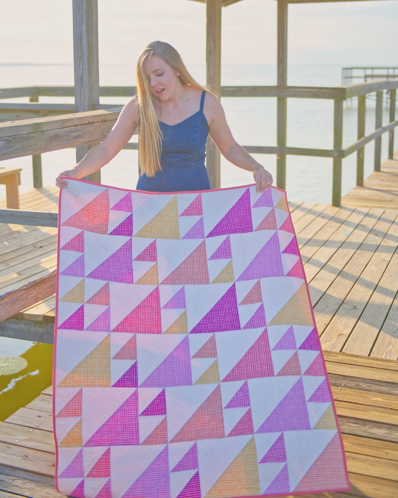 May include: A colorful quilt with a geometric pattern of triangles in shades of pink, orange, and white. The quilt has a pink border and is held up by a person on a wooden dock.