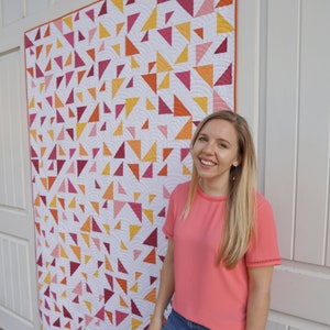 May include: A woman stands in front of a white wall with a large quilt hanging behind her. The quilt is made of white fabric with a pattern of colorful triangles in shades of pink, red, orange, and yellow.