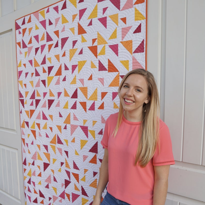 May include: A woman stands in front of a white wall with a large quilt hanging behind her. The quilt is made of white fabric with a pattern of colorful triangles in shades of pink, red, orange, and yellow.