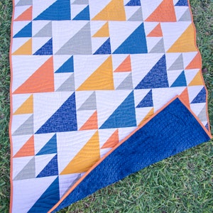 May include: A geometric quilt featuring a pattern of triangles in shades of blue, orange, yellow, and gray on a white background. The quilt has an orange border and a navy blue textured backing. The quilt is lying on green grass.