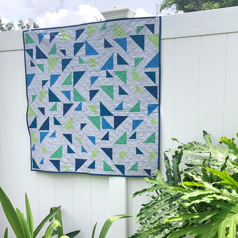 May include: A white quilt with a geometric pattern of blue and green triangles. The quilt is hanging on a white fence.