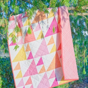 May include: A colorful quilt with a geometric triangle pattern in shades of pink, yellow, and orange, set against a white background. The quilt is trimmed with a solid pink border and a textured pink backing. The quilt is hanging on a tree branch.