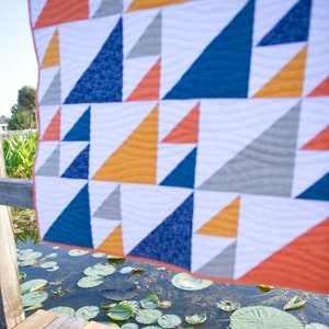 May include: A geometric quilt with a repeating triangle pattern in shades of orange, navy blue, gold, and gray on a white background. The quilt is hanging near a body of water with lily pads. The quilt's edge is orange.