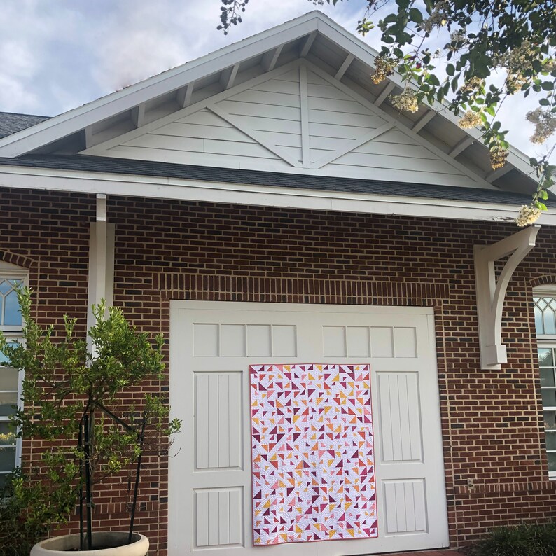 May include: A white brick building with a garage door. A pink, orange, and white quilt is hanging on the garage door.