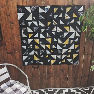 May include: A black and white quilt with yellow triangles. The quilt is made of fabric scraps and is hanging on a wooden wall.