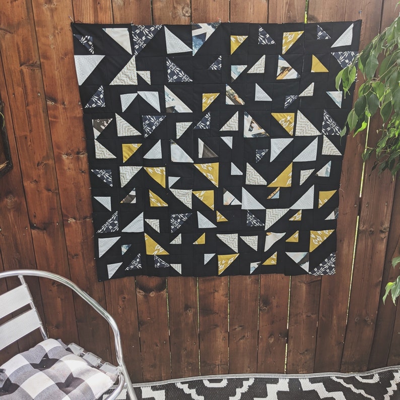 May include: A black and white quilt with yellow triangles. The quilt is made of fabric scraps and is hanging on a wooden wall.