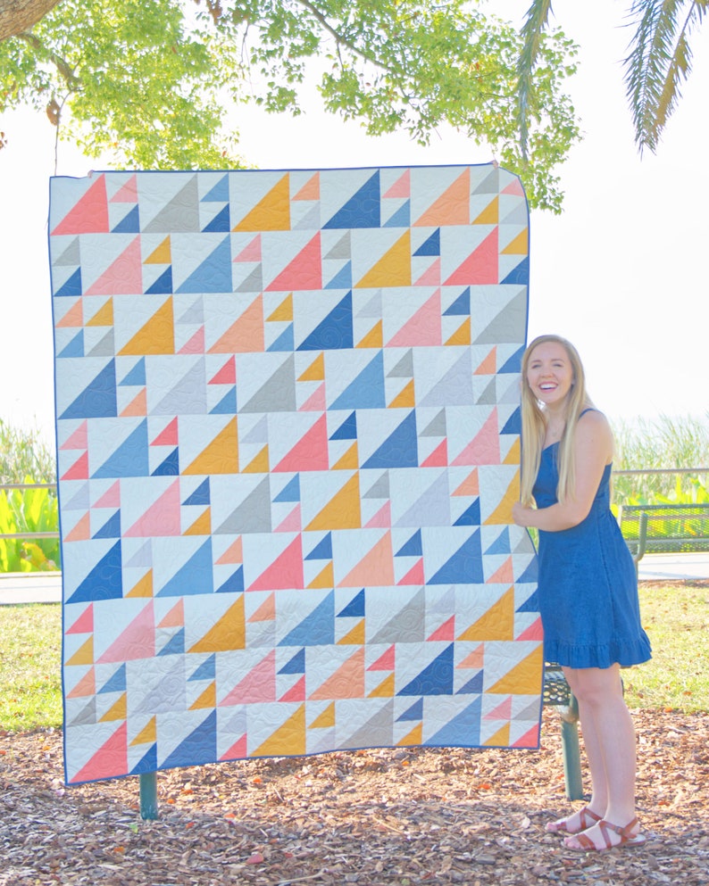 May include: A large, colorful quilt with a geometric triangle pattern in shades of blue, pink, yellow, and gray. The quilt is displayed outdoors, showcasing its design and craftsmanship. The quilt is a decorative home textile.
