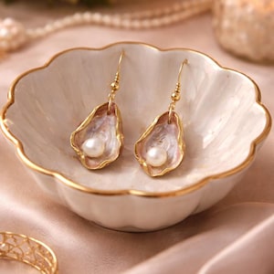 May include: A pair of gold-toned dangle earrings, each featuring a pearl nestled within a shell-shaped design. The shells have a pink and white interior with gold edging. The earrings rest in a white, scalloped dish with gold trim.
