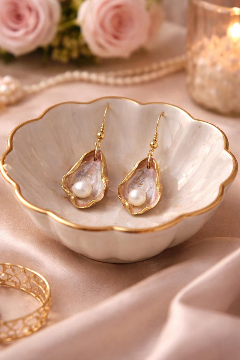 May include: A pair of gold-toned dangle earrings, each featuring a pearl nestled within a shell-shaped design. The shells have a pink and white interior with gold edging. The earrings rest in a white, scalloped dish with gold trim.