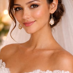 May include: A bride wearing a white off-the-shoulder lace wedding dress and veil. She is wearing gold earrings with a pearl-like center. Her hair is styled in an updo with loose curls framing her face. She has a soft, natural makeup look.
