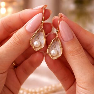 May include: Gold-tone oyster shell earrings with a white interior and a single pearl. The earrings are held up by a person. The earrings have a delicate, elegant design, perfect for special occasions.