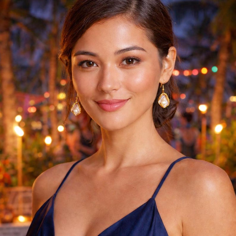 May include: A woman wearing gold drop earrings with a pear-shaped pearl and a dark blue spaghetti strap dress. The earrings have a gold frame. The background is blurred with lights and palm trees.