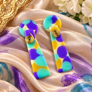 May include: A pair of dangle earrings featuring a circular top piece and a rectangular bottom piece, both decorated with a multi-colored pattern of purple, teal, and yellow circles. Each earring includes a gold-toned wire spiral accent.