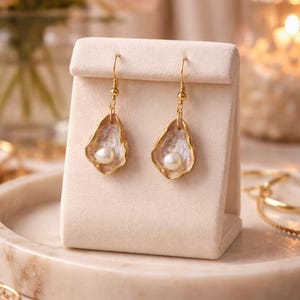 May include: A pair of gold-toned earrings featuring a pearl nestled within a shell-shaped design. The earrings are displayed on a cream-colored stand, with a soft-focus background of flowers and candles. The earrings are a delicate piece of jewelry.