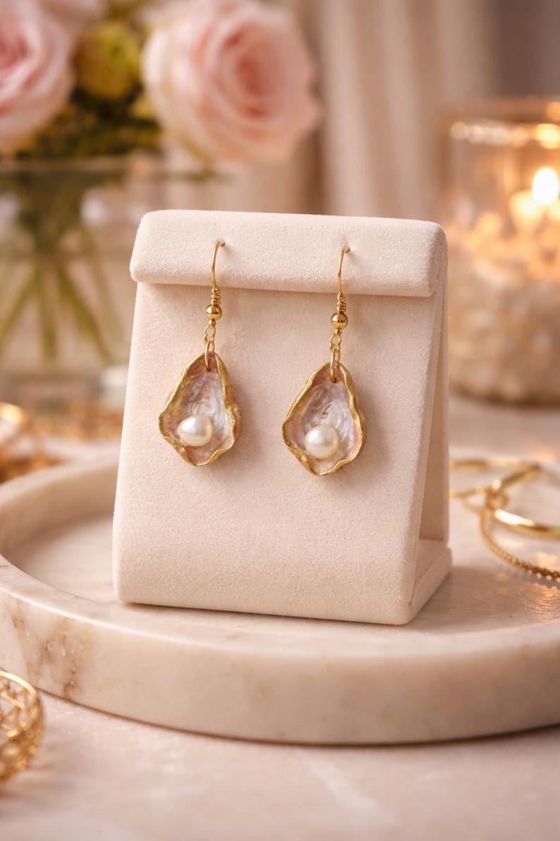 May include: A pair of gold-toned earrings featuring a pearl nestled within a shell-shaped design. The earrings are displayed on a cream-colored stand, with a soft-focus background of flowers and candles. The earrings are a delicate piece of jewelry.