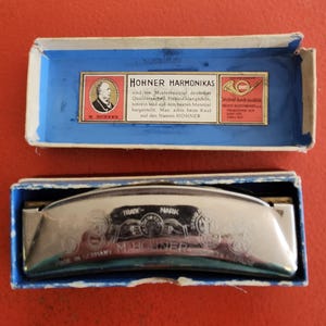 May include: A vintage Hohner harmonica in its original blue and white box. The harmonica is silver-toned with the words "TRADE MARK" and "MADE IN GERMANY M. HOHNER" engraved on it. The box lid has text in German.